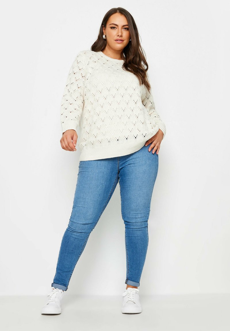 Elfenbeinfarbener, gestrickter Pullover mit Rautenmuster, Rundhalsausschnitt und langen Ärmeln, kombiniert mit hellblauen Skinny-Jeans und weißen Sneakers.