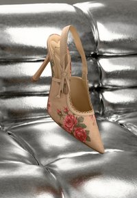 Zapato stiletto con tira trasera en ante beige con estampado floral rosa, punta picuda y ribete de encaje. Fondo plateado brillante.