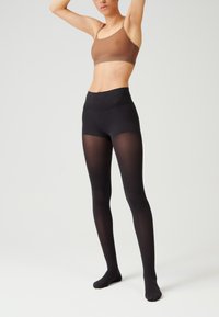 Bralette en bronze avec des fines bretelles et une bande avec logo, associé à des collants noirs transparents. Les collants ont une texture lisse et un design ajusté.