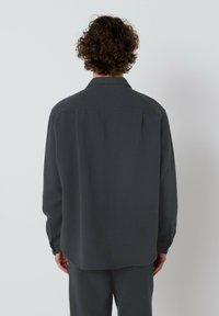 Oseba s kodrastimi lasmi, oblečena v dolg rokav temno sivo srajco in ustrezne hlače, stoji obrnjena proti enobarvni beli steni.