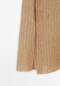 Tissu en lin marron avec fines rayures blanches verticales, montrant une fente latérale et une texture douce.