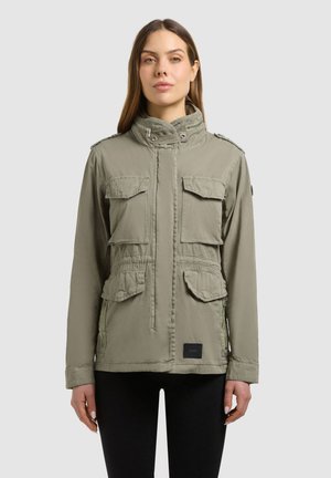 Žena v lehkém olivově zeleném vojenském kabátu se čtyřmi klopovými kapsami stojí na jednoduchém světle šedém pozadí.