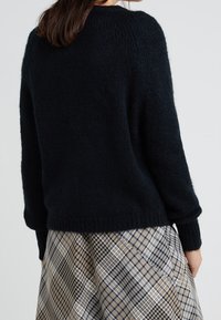 Maglione nero lavorato a maglia con una vestibilità ampia, scollatura rotonda e maniche leggermente affusolate, abbinato a una gonna a quadri nei colori beige e blu navy.