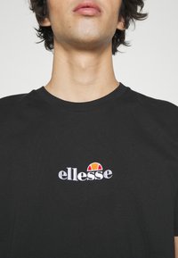 Sort bomulds T-shirt med rund halsudskæring og broderet "ellesse" logo i hvidt med en multifarvet halvcirkeldetalje.