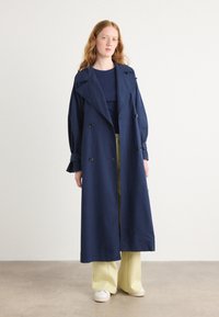 Manteau long bleu marine à double boutonnage, avec un large col et des manches amples, associé à un pantalon large beige et des baskets blanches.