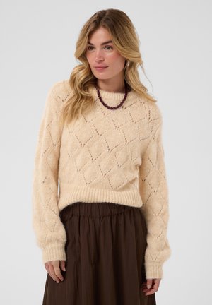 Femme portant un pull crème en maille à motif losange, une jupe brune et un collier de perles violettes, debout devant un fond uni.