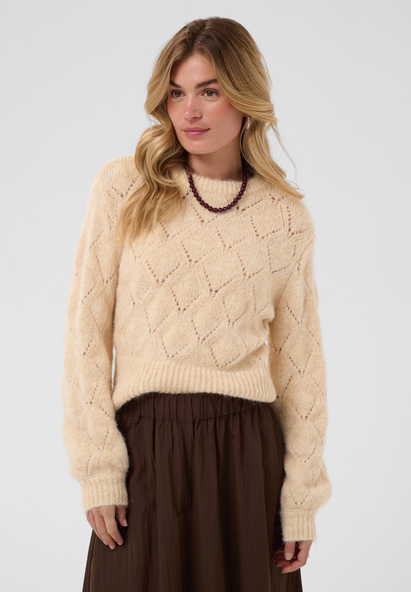 Femme portant un pull crème en maille à motif losange, une jupe brune et un collier de perles violettes, debout devant un fond uni.