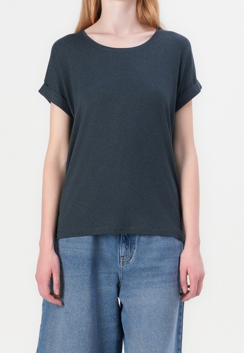 Personne portant un t-shirt gris foncé à manches courtes avec les manches retroussées et un jean taille haute bleu clair sur un fond uni.