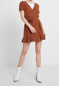 Robe portefeuille à pois marron avec un ourlet à volants, de courtes manches bouffantes et un détail de taille à nouer, associée à des bottines blanches.