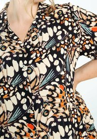 Vrouw die een overhemd draagt met knopen, bedrukt met een kleurrijk vlinder vleugelpatroon in beige, zwart, oranje en blauwtinten op een witte achtergrond.