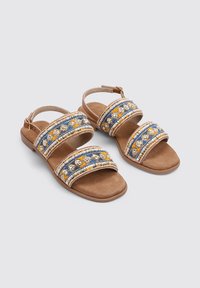 Sandalias marrones con tres tiras de cuentas que presentan patrones en azul, amarillo y blanco. Plantilla de ante suave con suela de goma plana. Correa ajustable en el tobillo.