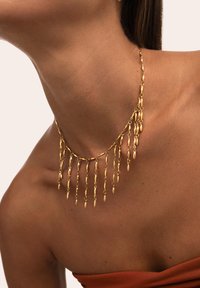 Collana d'oro con elementi pendenti, link delicati e un design curvato che poggia sulla clavicola, abbinata a un top senza spalline.