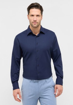 Mand iført en mørkeblå skjorte med lange ærmer og knapper samt lyseblå bukser, stående op ad en ensfarvet lys baggrund.