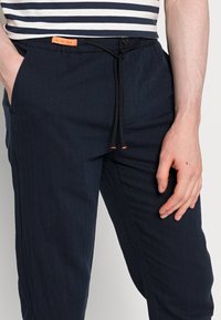 Marineblauwe broek met een trekkoord in de taille en oranje accenten, uitgevoerd met zijzakken en een gestructureerde stof. Het ontwerp is casual en ontspannen.