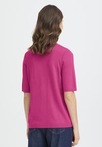 Fukszia rövid ujjú felső sima kötött anyagból, laza szabással, egyszerű gallérral és egyenes szegéllyel, sötét farmerrel párosítva.