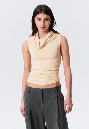 Jeune femme aux cheveux foncés portant un haut beige sans manches à encolure drapée et un pantalon gris à fines rayures, debout devant un fond uni.