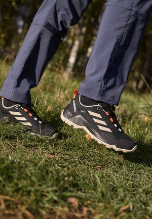 Personne portant des chaussures de randonnée Adidas noires et beiges marchant sur l'herbe en extérieur avec un pantalon bleu visible.