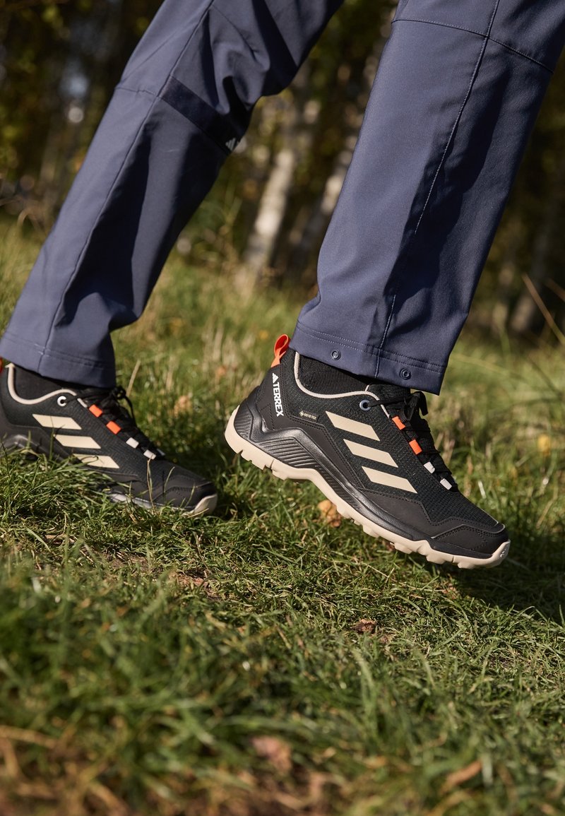 Personne portant des chaussures de randonnée Adidas noires et beiges marchant sur l'herbe en extérieur avec un pantalon bleu visible.
