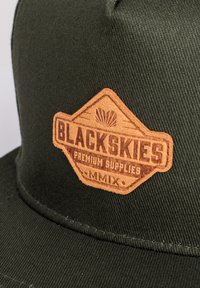 Cappellino verde in tessuto texture, con una patch in pelle marrone. La patch mostra la scritta "BLACKSKIES PREMIUM SUPPLIES" e "MMIX" in testo marrone.