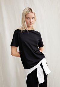 Zwarte t-shirt met korte mouwen en een ronde hals, gemaakt van zachte stof. Draag het over een witte overhemden die op de taille is geknoopt.