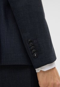 Navy blazer mouw met fijn gestructureerde stof, voorzien van vier zwarte knopen en een witte shirtmanchet die eronder uitkomt.