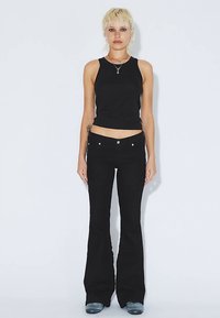 Černý tílko bez rukávů a černé úzké džíny s rozšířenými nohavicemi. Tílko má hladkou texturu; džíny jsou opatřeny předními kapsami a knoflíkovým detailem.