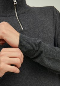 Maglione grigio scuro a maglia con colletto alto, chiusura con zip argentata e polsini a costine. Il tessuto sembra morbido e texture.