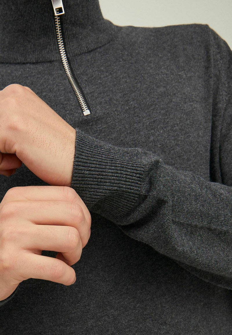 Maglione grigio scuro a maglia con colletto alto, chiusura con zip argentata e polsini a costine. Il tessuto sembra morbido e texture.