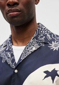 Μπλούζα κοντό μανίκι σε ναυτικό μπλε χρώμα με λευκά λουλουδάτα και αφηρημένα σχέδια, διαθέτει κομμένο γιακά και κουμπί σε μια μόνο θέση, φορεμένη πάνω από λευκή υποκάμισο.