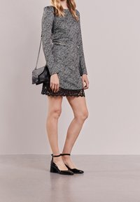 Robe grise texturée avec des manches longues et un ourlet en dentelle, assortie à des escarpins en vernis noir. Sac bandoulière noir avec des détails en métal argenté.