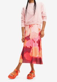 Rosa gekürztes Sweatshirt mit einem erhabenen Logo, kombiniert mit einem langen, mehrfarbigen Rock mit abstrakten Mustern und orangefarbenen Plateausandalen.