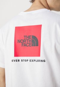 Hvitt bomull t-skjorte med et stort rødt kvadratisk grafikk med "THE NORTH FACE" og "NEVER STOP EXPLORING" i svart skrift.