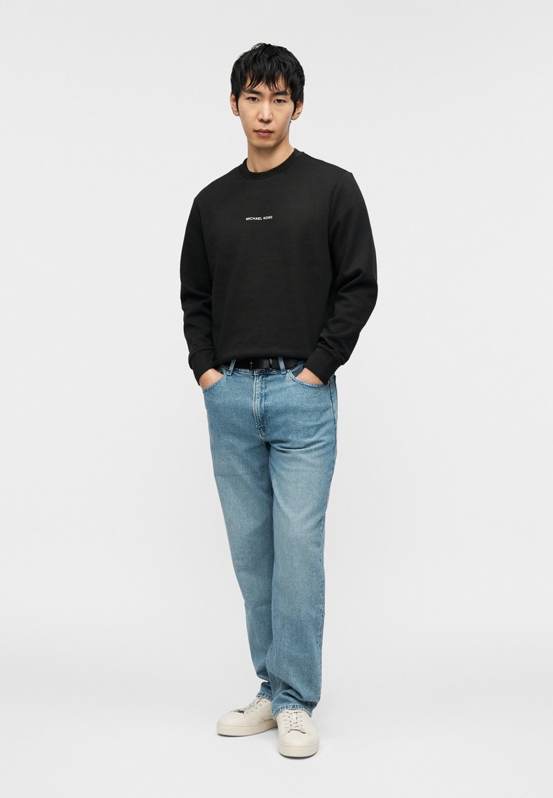 Μαύρο φούτερ με στρογγυλή λαιμόκοψη και κείμενο στο μπροστινό μέρος, συνδυασμένο με ανοιχτό μπλε τζιν και κρεμ αθλητικά παπούτσια. Άνετη εφαρμογή με ομαλή υφή.