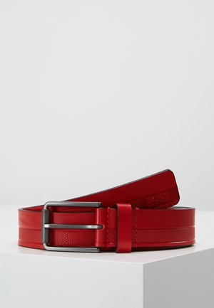 Ceinture en cuir rouge avec motif texturé et boucle en métal, enroulée sur une surface blanche.