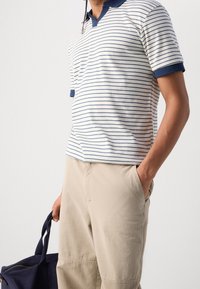 Pruhovaná polokošile s námořnickým límcem a rukávy, z bavlněného materiálu. Béžové kalhoty, ruka v kapse, držící námořnickou tašku.