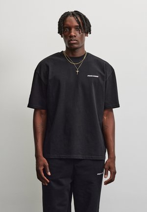 Jeune homme aux cheveux tressés portant un t-shirt et un pantalon noirs Pegador, debout devant un fond gris clair uni.