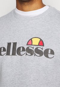 Hombre que lleva un sudadera gris claro con un logo negro de "ellesse" y un diseño de semicírculo rojo y amarillo en el pecho.