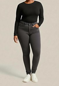 Femme portant un haut noir à manches longues, un jean skinny gris foncé et des baskets blanches, debout avec une main dans la poche devant un fond uni.