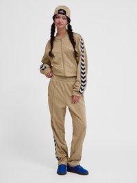 Noor naine punutud juustega, kandes beeži dressi meresiniste ševroontriipudega, tagurpidi beeži nokamütsi ja erkrohelisi tossusid, seistes lõdvestunult.