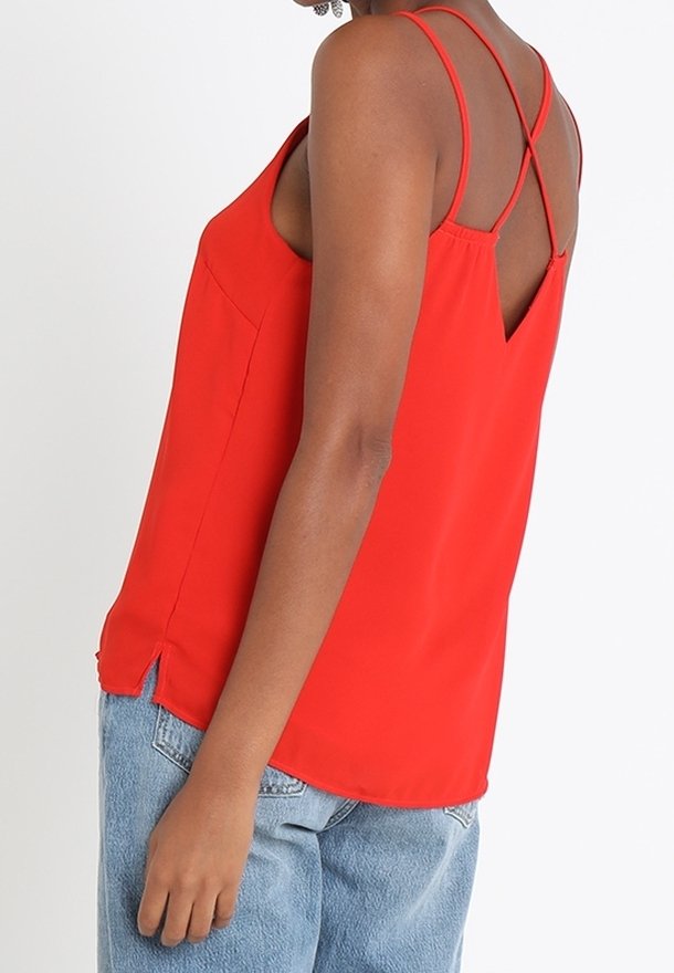 Blouse rouge sans manches avec un dos en V, de fines bretelles croisées, des fentes latérales et une texture lisse, associée à un jean bleu clair.