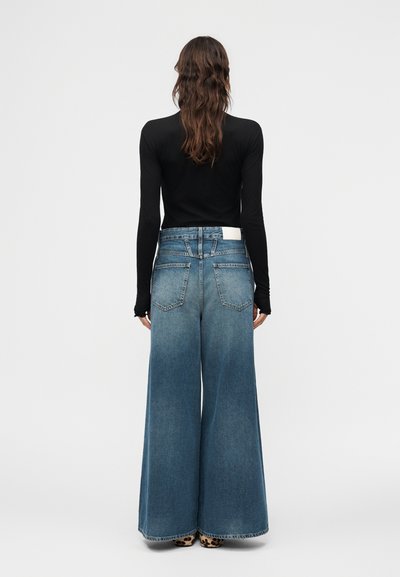 Γυναίκα με μακριά κυματιστά καστανά μαλλιά φοράει εφαρμοστό μακρυμάνικο μαύρο τοπ και φαρδιά ξεβαμμένα μπλε τζιν, στέκεται μπροστά σε απλό φόντο.