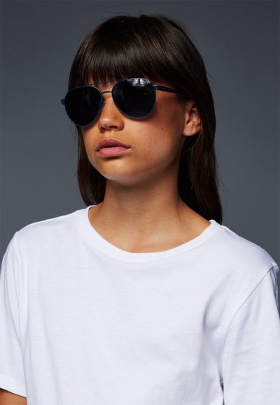 Óculos de sol de armação redonda com lentes escuras e uma estrutura metálica azul. Usados com uma t-shirt branca simples contra um fundo neutro.