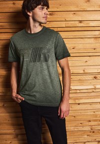 Olivově zelené bavlněné tričko s texturovaným povrchem, se silným grafickým potiskem v černé. Nosené s černými kalhotami před dřevěným pozadím.