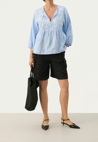 Blusa azzurra chiara ricamata con scollo a V e maniche a tre quarti, abbinata a pantaloni cargo neri e décolleté nere a punta. Borsa a tracolla intrecciata nera.