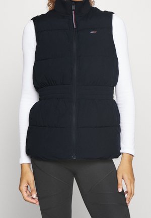 Personne portant un gilet sans manches noir rembourré avec fermeture éclair par-dessus un haut blanc à manches longues et des leggings foncés, debout devant un fond uni.
