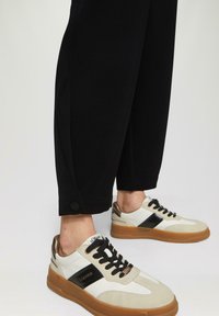 Witte en zwarte sneaker met een tan zool, voorzien van vetersluiting, zijstrepen en metallic accenten. Gecombineerd met zwarte tapered broek.