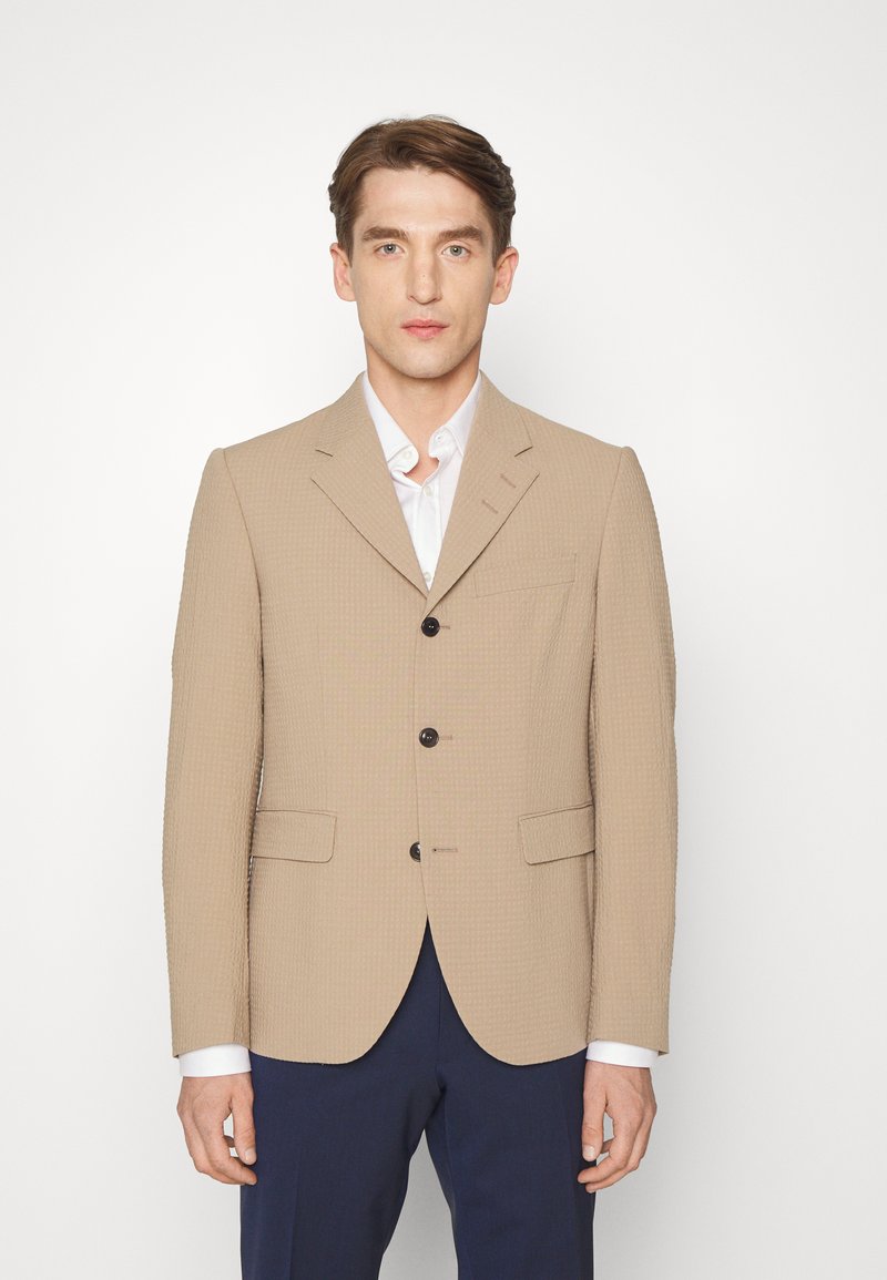 Blazer texturizado bege com lapelas em notched, dois bolsos frontais, fecho de três botões e botões escuros em contraste, usado sobre uma camisa branca.