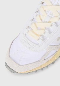 Primer plano de la puntera de una zapatilla blanca y beige con suela texturizada y cordones crema sobre un fondo claro.