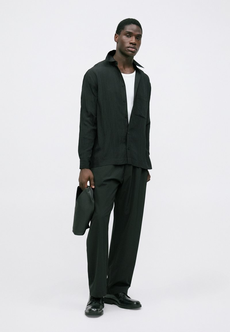 Mand iført sort skjorte over hvid top, sorte vide bukser, sorte sko, holder en sort læder clutch mod en ensfarvet baggrund.