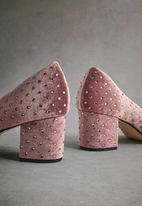 Vaaleanpunaiset samettiset korokepohjaiset kengät, joissa on ripoteltuja strassikivitä, terävät varpaat ja teksturoitu pinta. Kannan korkeus on noin 7,5 cm.